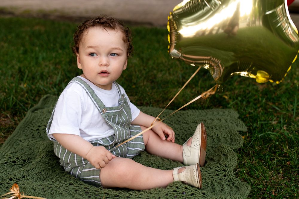 Baby boy in striped overalls sitting on a green blanket outdoors with a gold star balloon beside him.