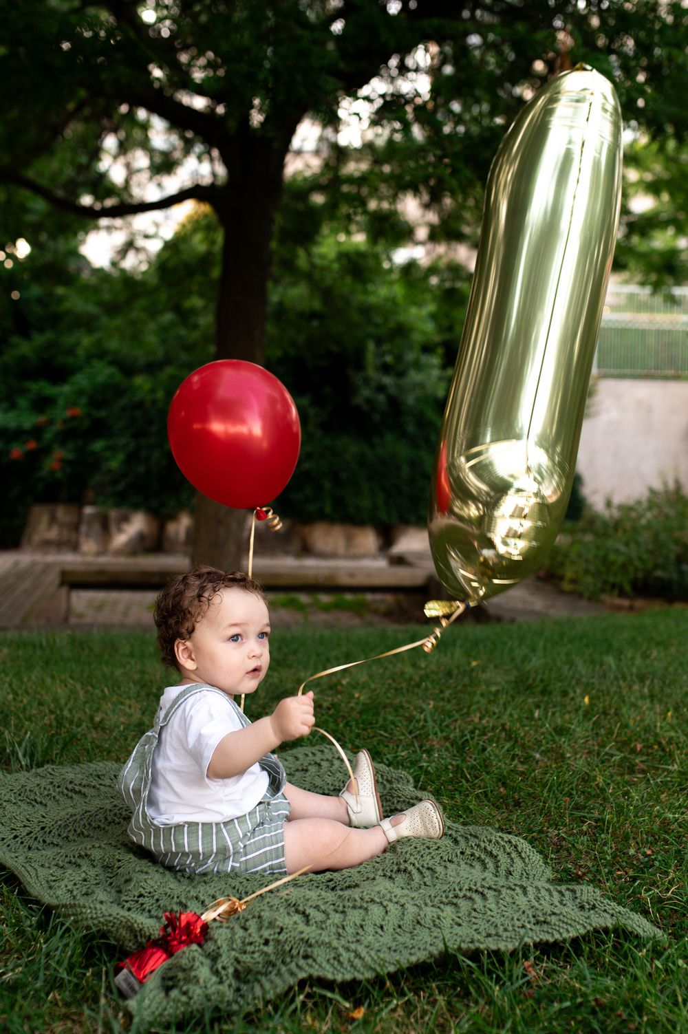 Baby boy sits on green blanket outdoors holding red and gold number one balloons for his first birthday.