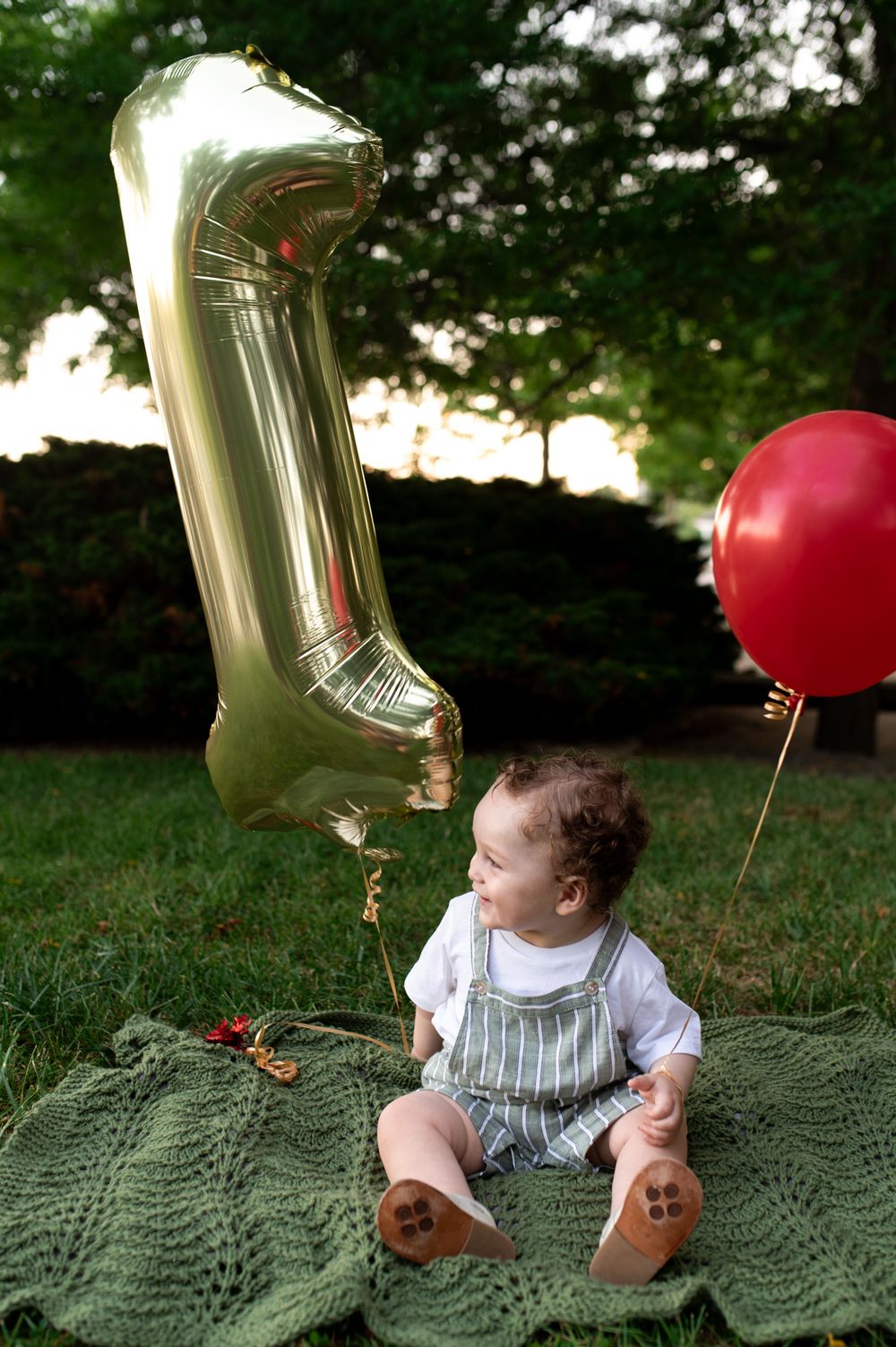 Baby boy sits on green blanket outdoors beside gold number one balloon and red balloon for first birthday.