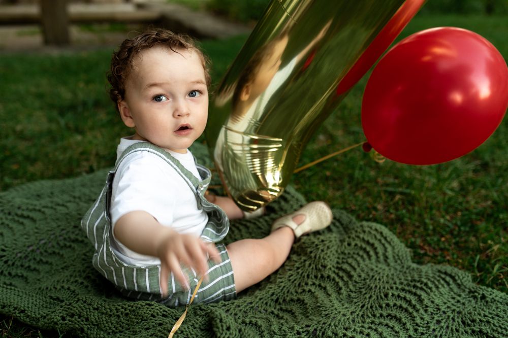 Adorable baby boy in suspenders sits on green blanket outdoors with gold number one and red balloon.