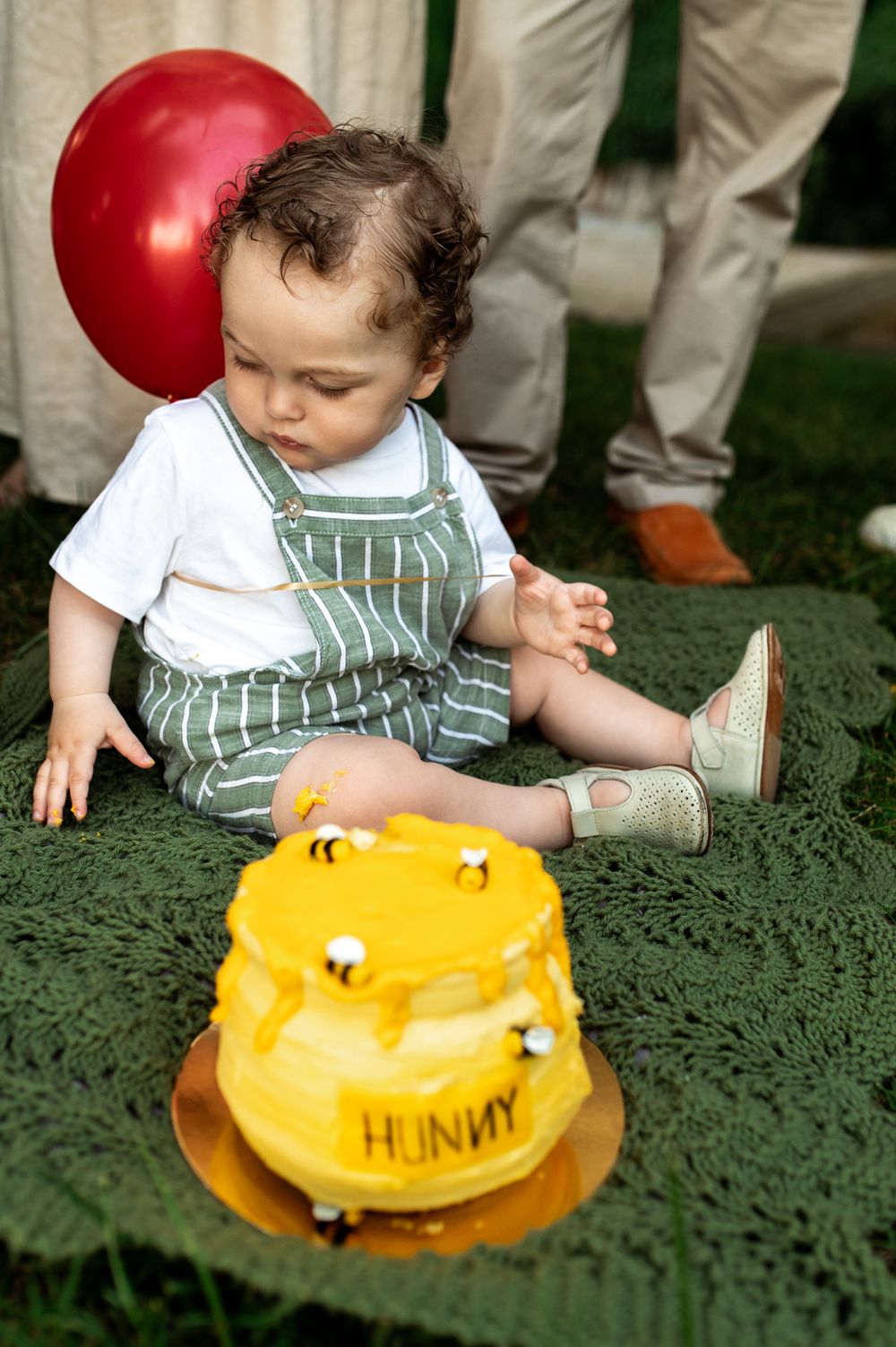Baby boy in striped overalls touching a yellow Winnie the Pooh Hunny pot smash cake at his first birthday party.