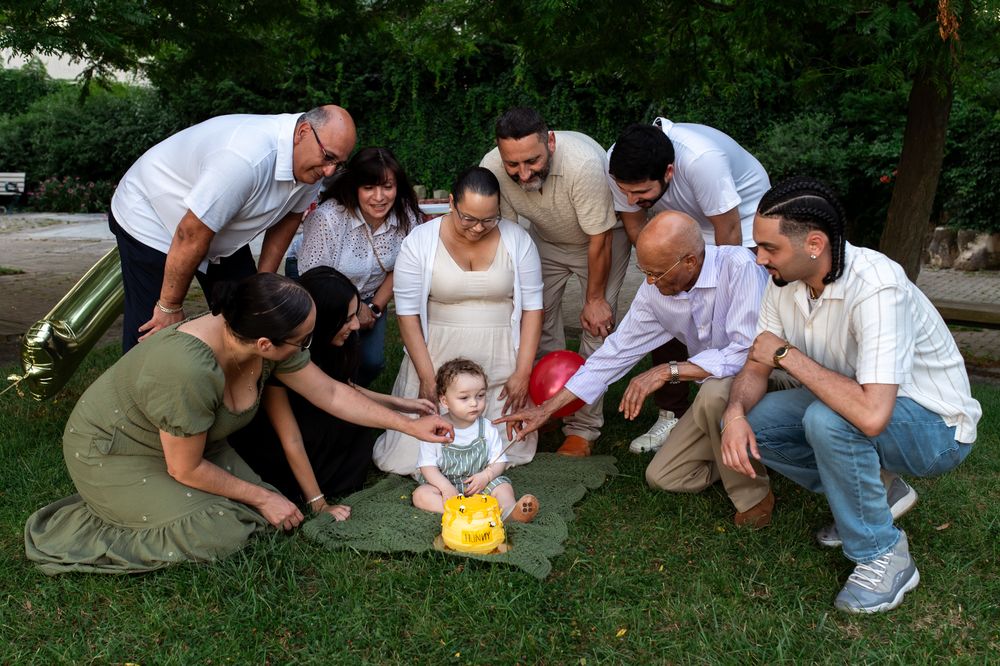Family gathering outdoors on green grass, celebrating baby's first birthday with a yellow piggy bank cake.