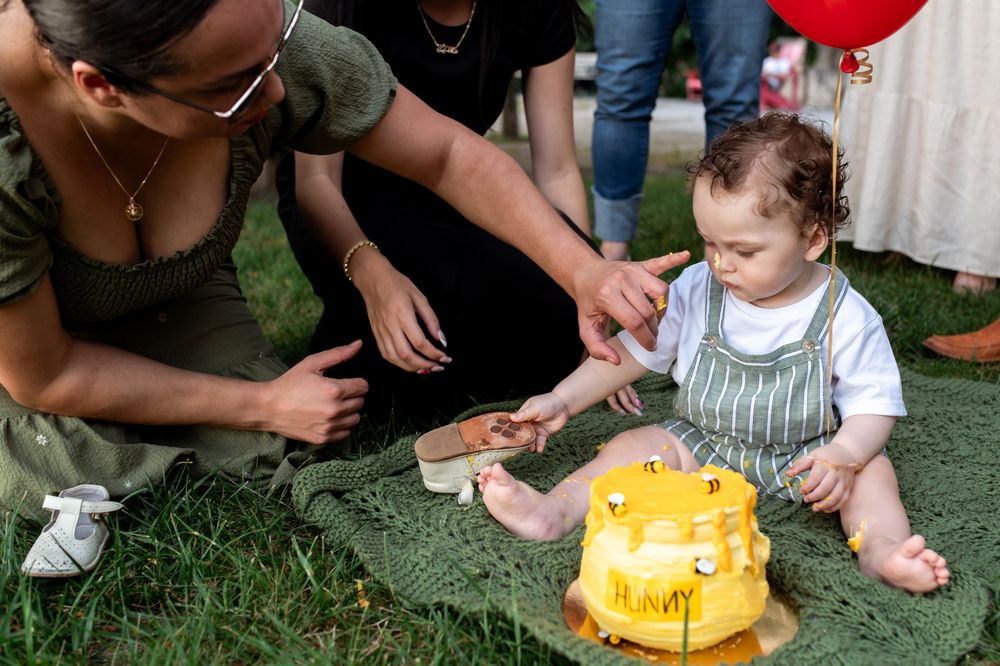Baby enjoys first birthday cake smash outdoors on grass while parents help celebrate the milestone moment.