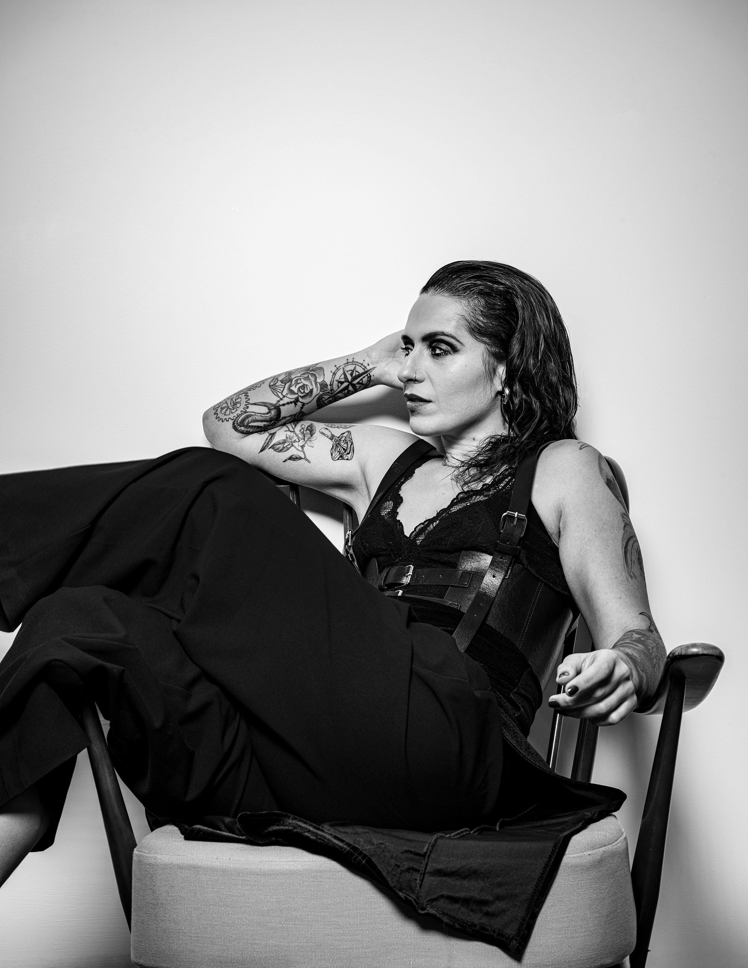 Black and white portrait of a tattooed woman reclining and drinking, seated in a wooden chair