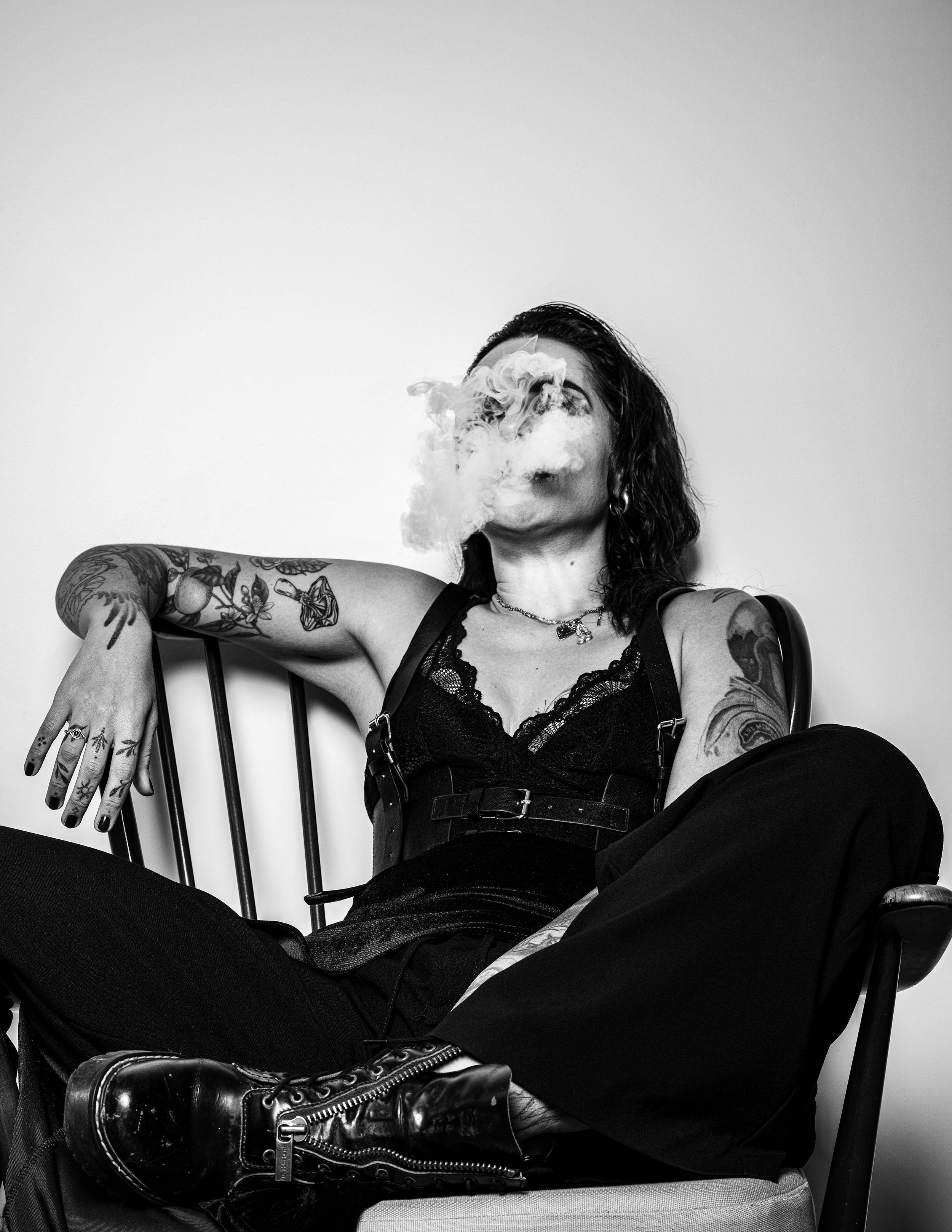 Black and white portrait of a tattooed woman exhaling smoke, dynamic motion, seated in a wooden chair