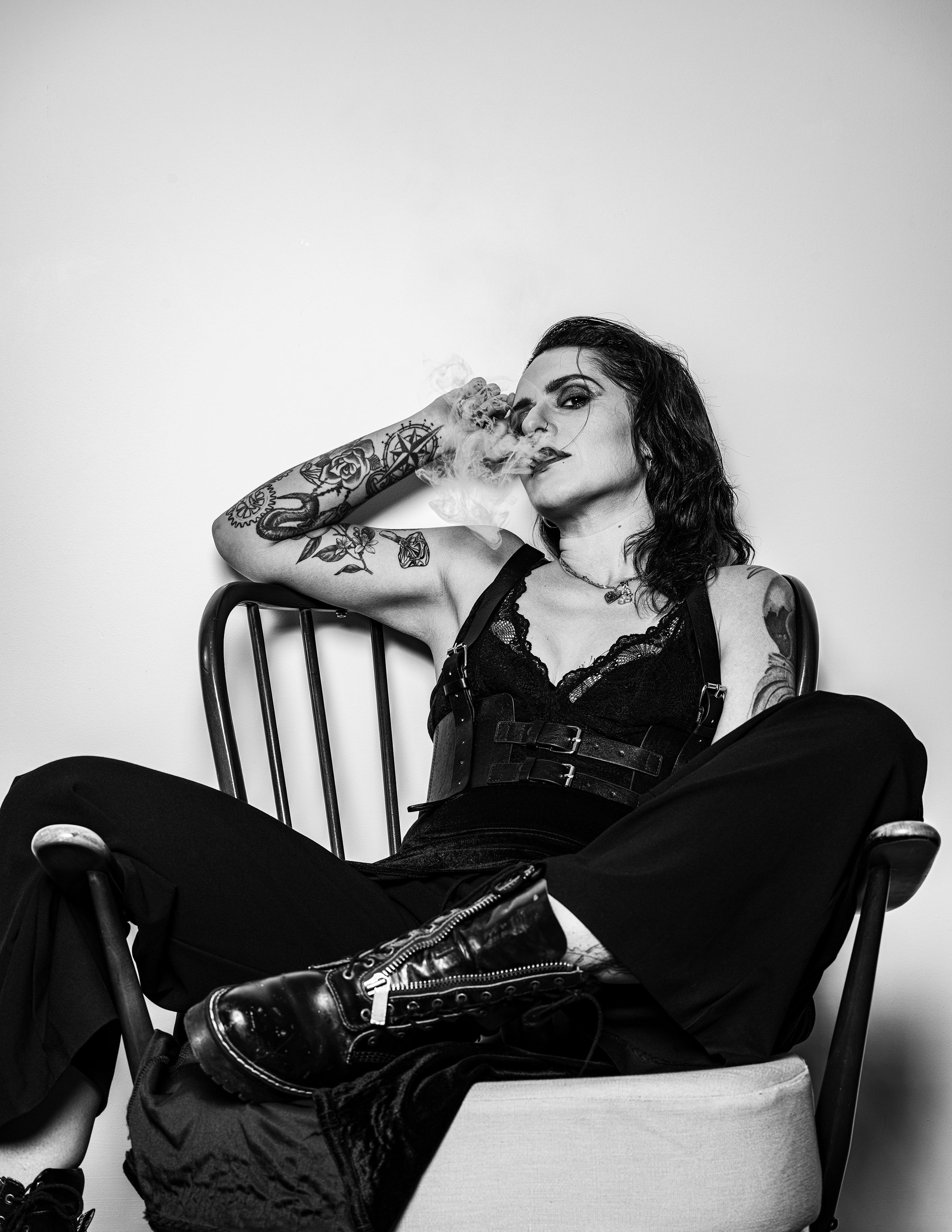 Black and white portrait of a tattooed woman smoking, seated in a wooden chair, moody editorial style