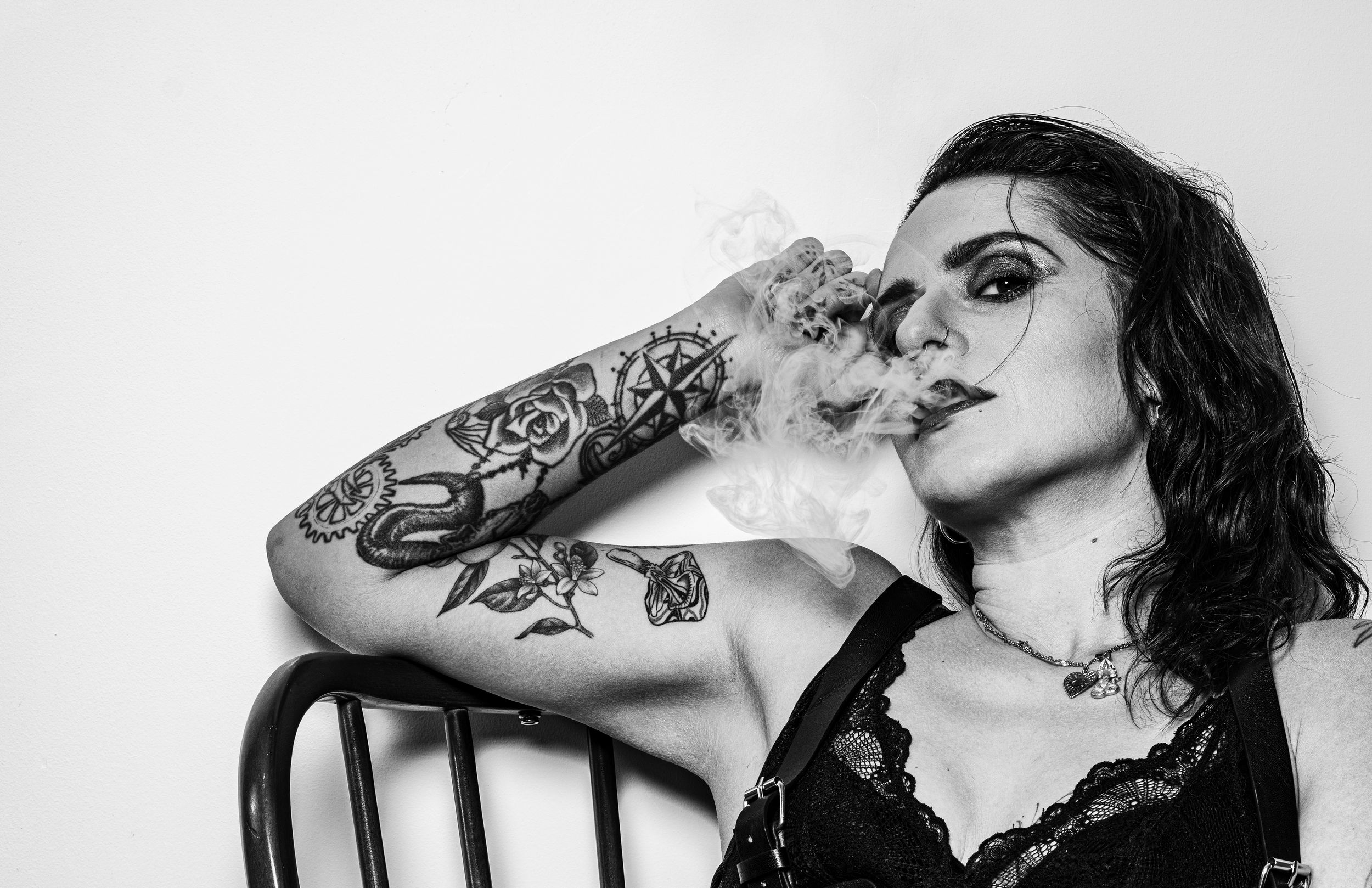 Black and white portrait of a tattooed woman smoking a cigarette in a lace top, seated in a wooden chair