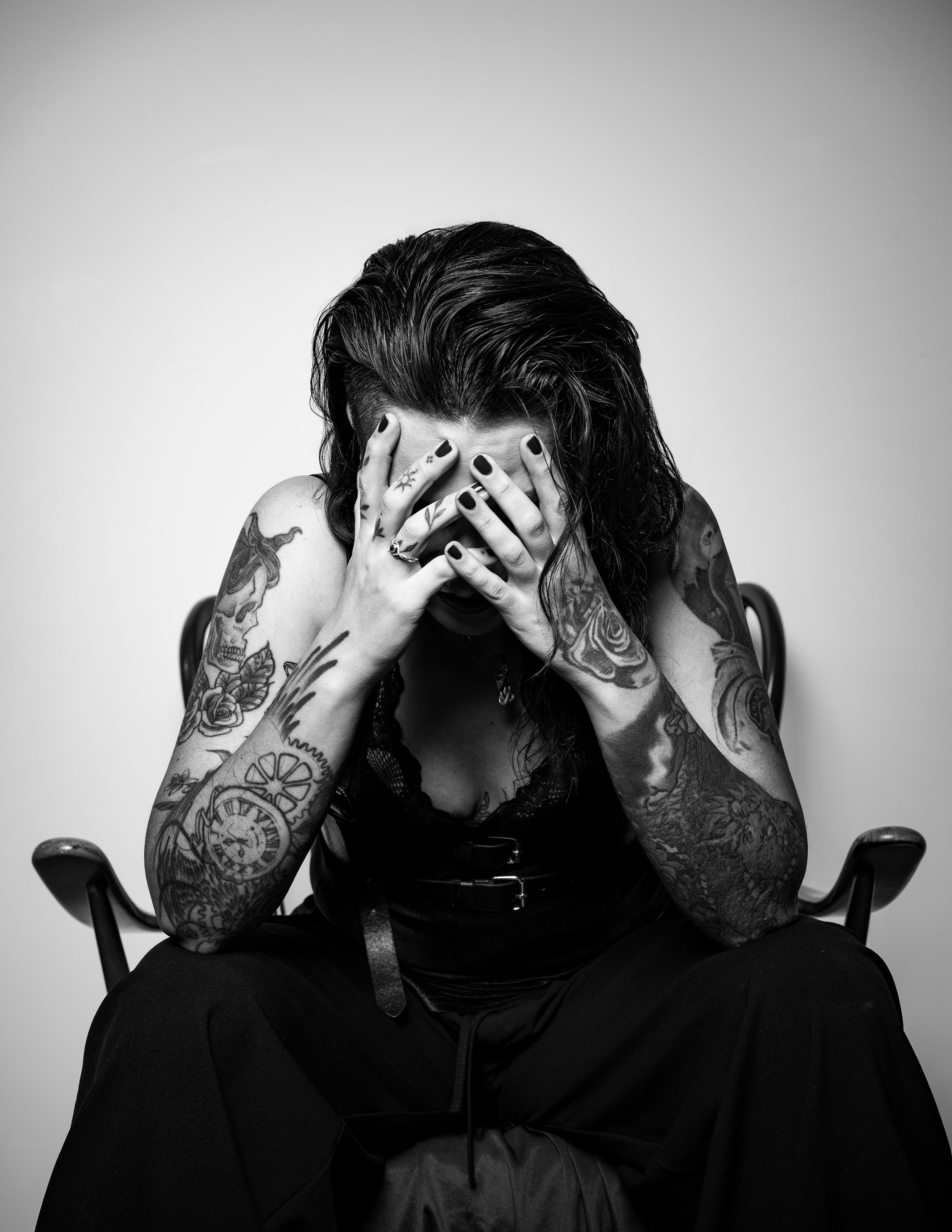 Black and white portrait of a tattooed woman covering her face with her hands, seated in a wooden chair, emotional editorial style