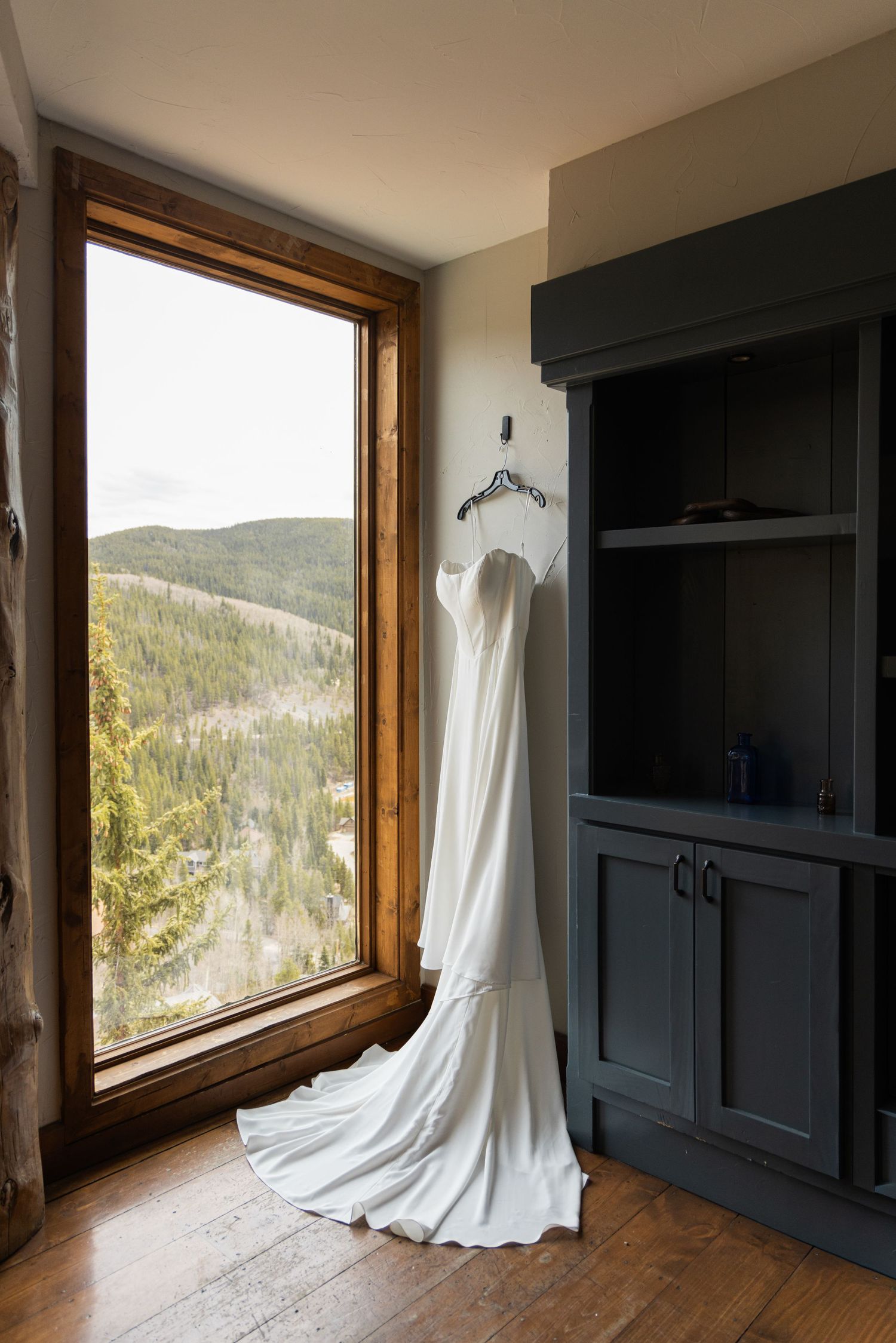 White wedding gown hanging on a hanger near a rustic wooden window with a scenic mountain forest view.