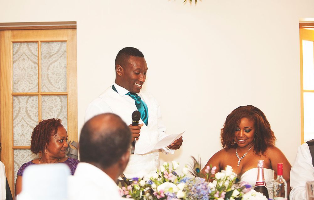 Groom giving a speech at wedding reception holding microphone, bride smiling beside him at head table. At Oaks farm wedding venue in Surrey.