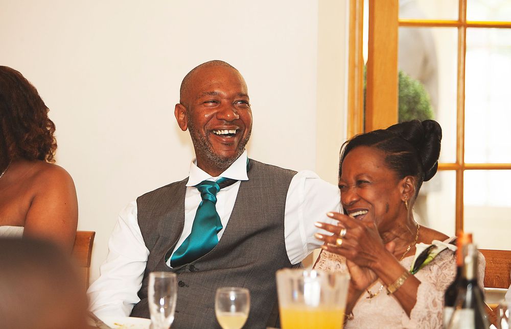 Laughing man in grey vest and teal tie seated at wedding reception table with smiling woman beside him. At Oaks farm wedding venue in Surrey