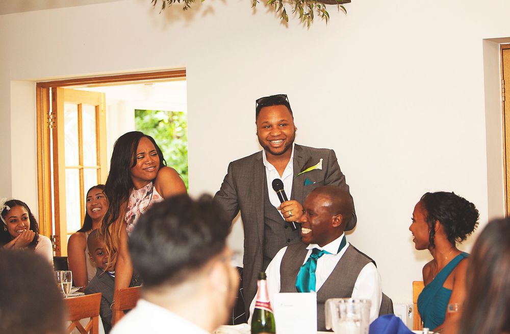 Man giving a speech at a wedding reception with guests smiling and listening in the background. At Oaks farm wedding venue in Surrey.