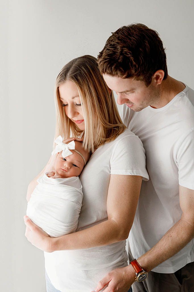 New parents in white shirts tenderly hold their swaddled newborn baby girl with a white bow headband.