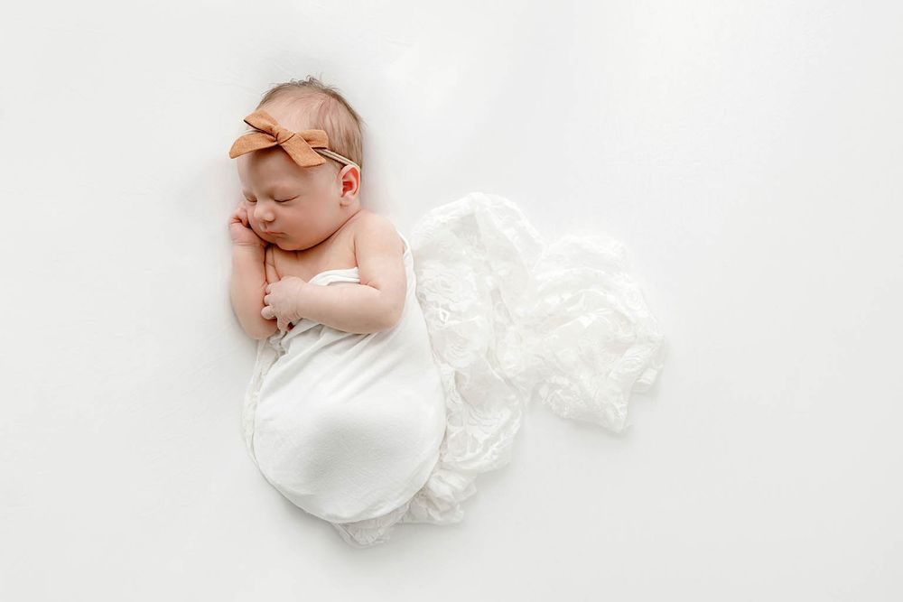Peaceful newborn baby swaddled in white wrap with orange bow headband on white background.