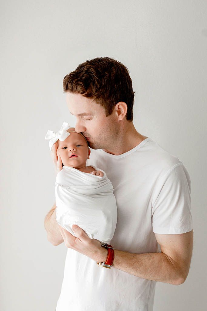 Father in white t-shirt tenderly kissing his swaddled newborn baby wearing a white bow headband.