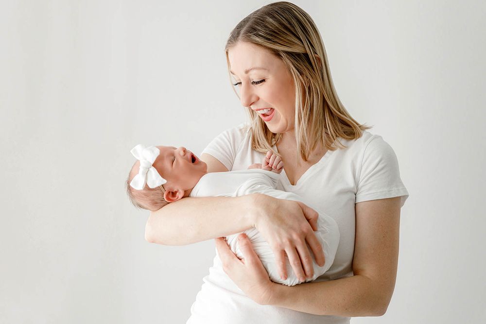 A smiling mother in a white shirt tenderly holds her newborn baby with a white bow headband against a light background.