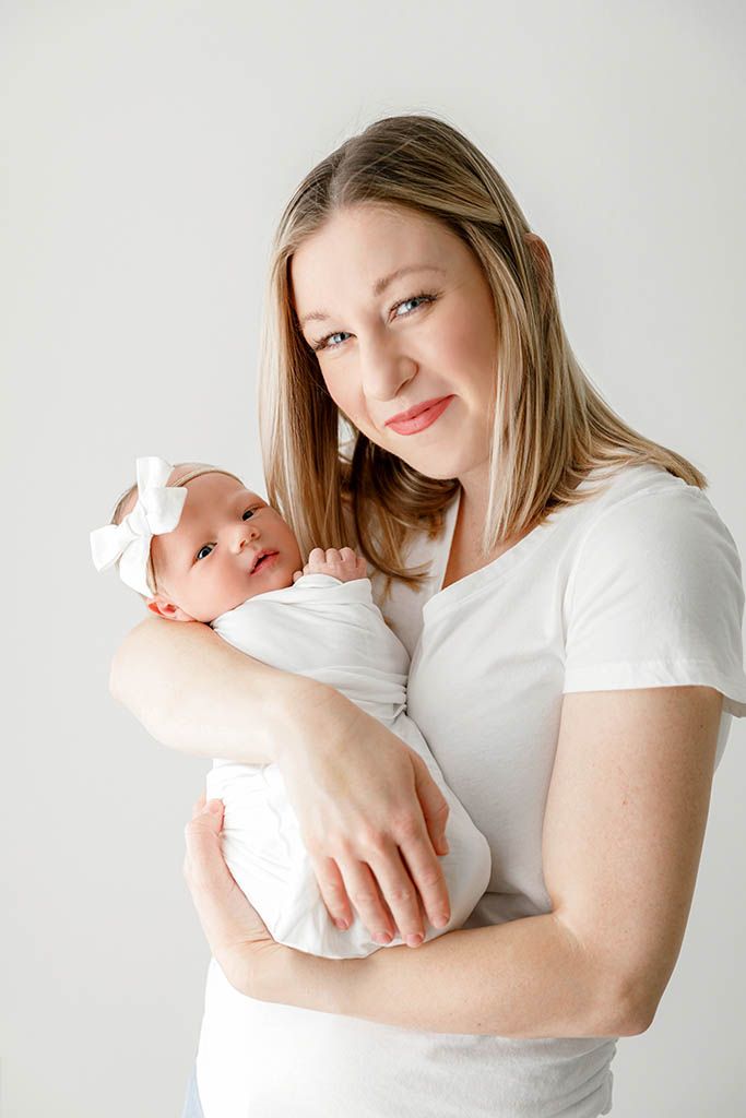 A smiling woman in a white shirt holds a swaddled newborn baby wearing a white bow headband against a light background.