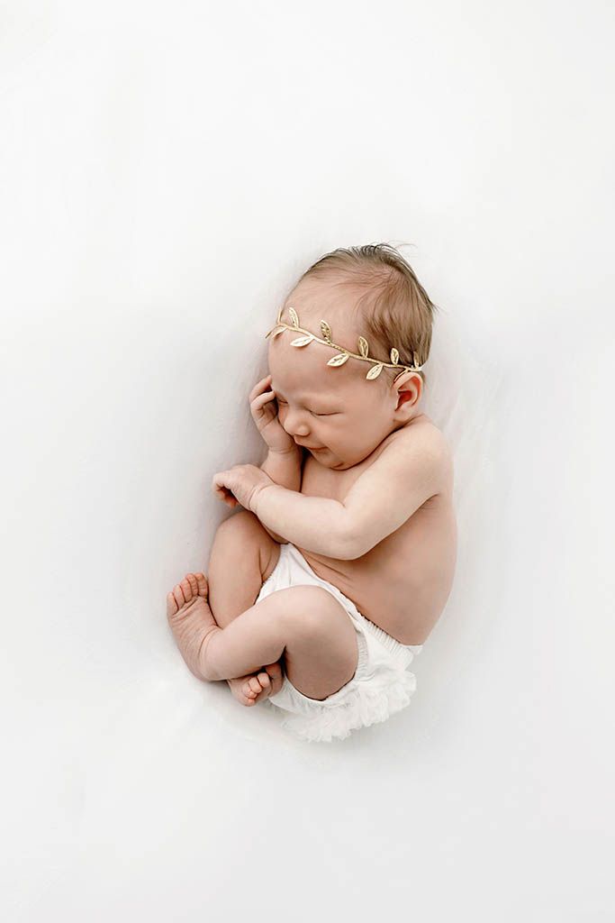 Sleeping newborn baby wearing a gold leaf headband, curled up on a white blanket in a peaceful pose.