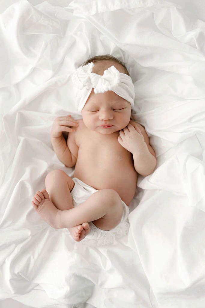 Peaceful sleeping newborn baby girl wearing white bow headband lying on soft white fabric.