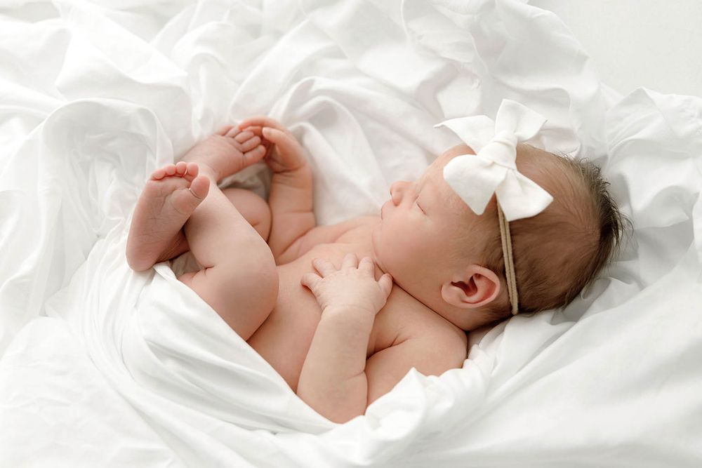 Serene newborn baby girl sleeping on white fabric wearing a white bow headband.