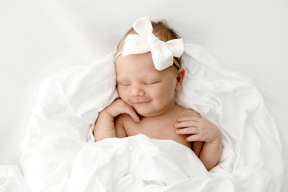 Smiling newborn baby sleeping peacefully wrapped in white blankets wearing a white bow headband.