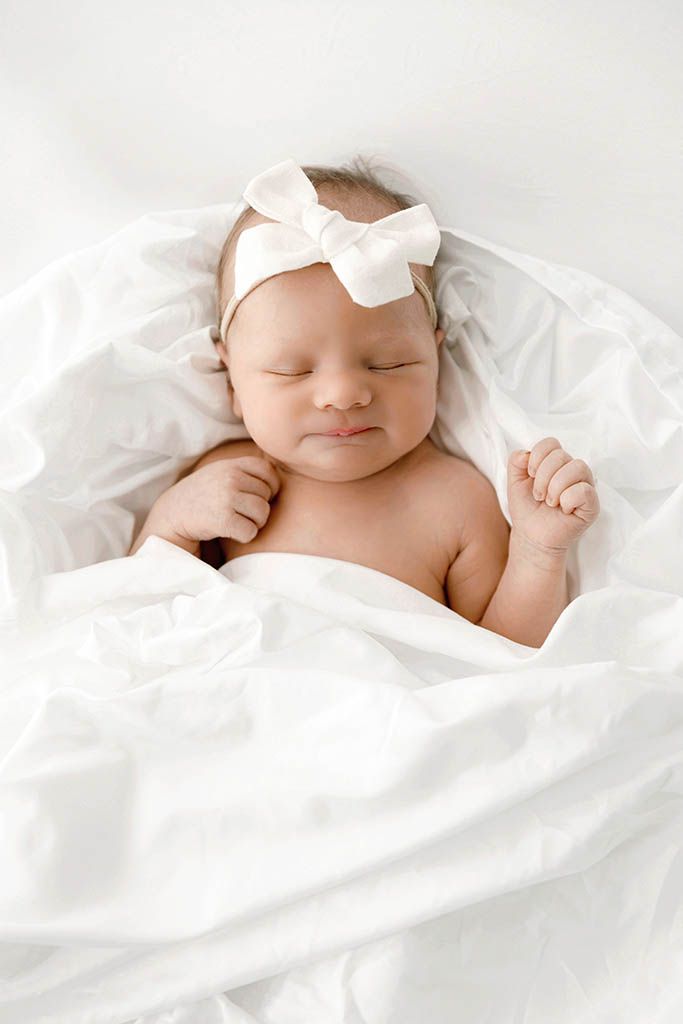 Newborn baby girl sleeping in white blankets wearing a white satin bow headband.