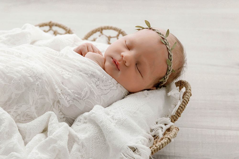 Newborn baby sleeping peacefully in a wicker basket, wrapped in white lace, wearing a delicate green leaf headband.