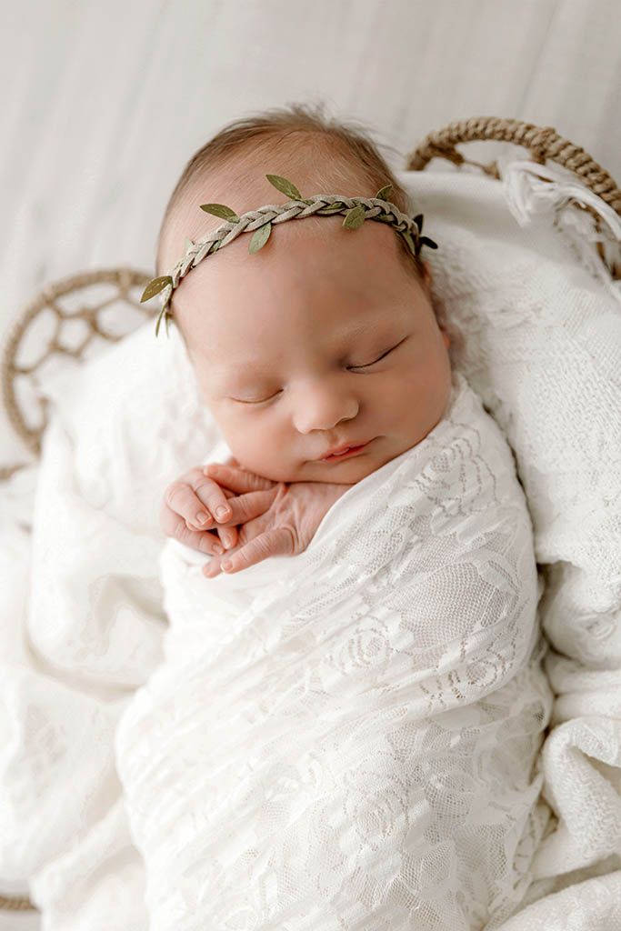 Sleeping newborn baby swaddled in white lace wrap wearing a delicate green leaf crown, resting peacefully in a wicker basket.