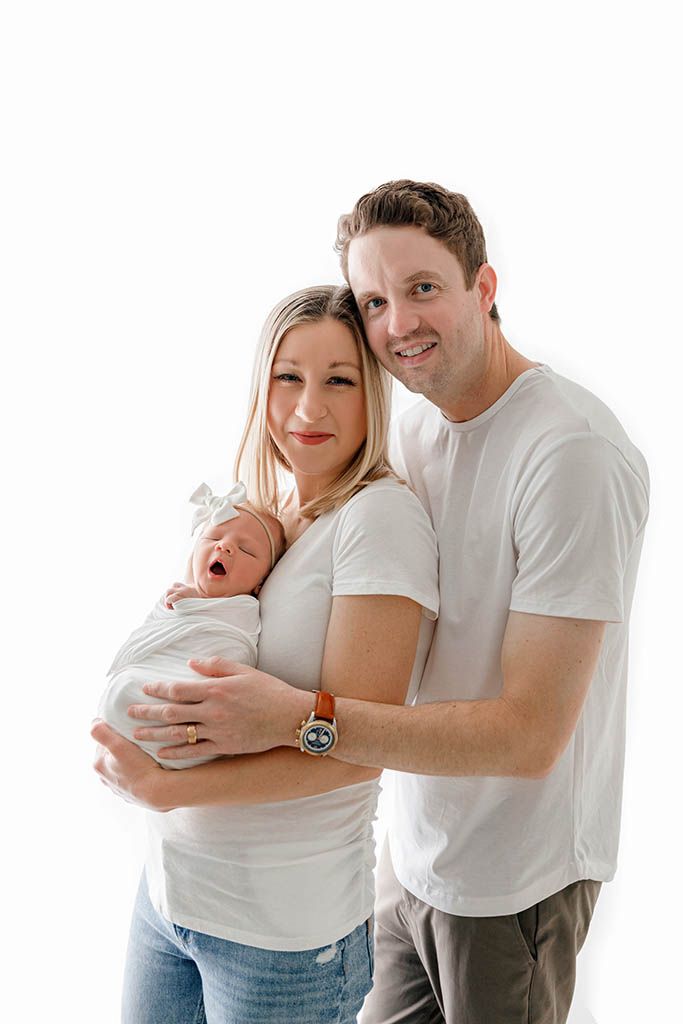 Happy parents in white shirts smile while holding their swaddled newborn baby against a white background.