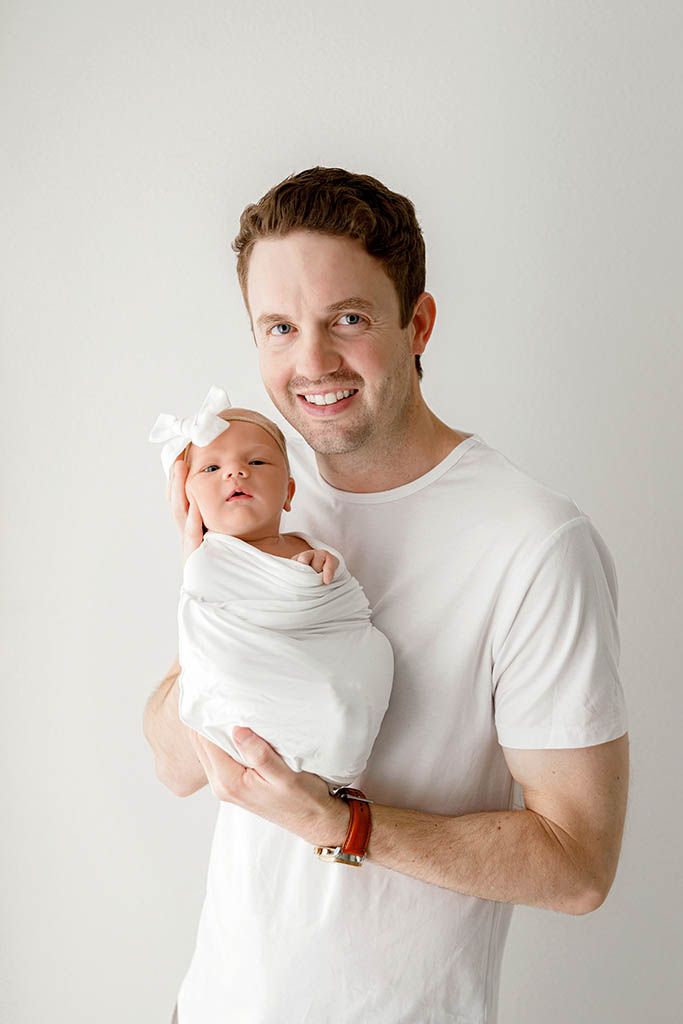 Smiling father in white t-shirt proudly holds his swaddled newborn baby girl with white bow headband.