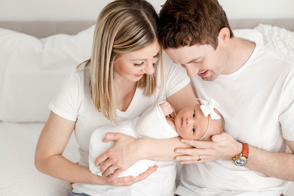 A couple in white shirts lovingly gazes at their swaddled newborn baby on a white bed.
