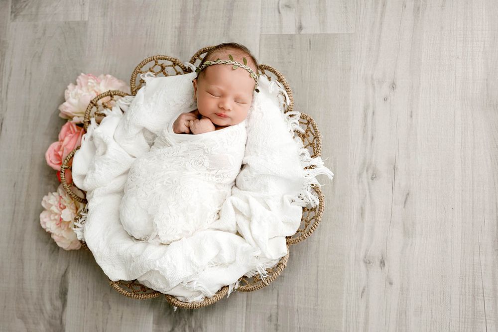 Newborn baby sleeping peacefully in a wicker basket with white wrap and pink flowers.
