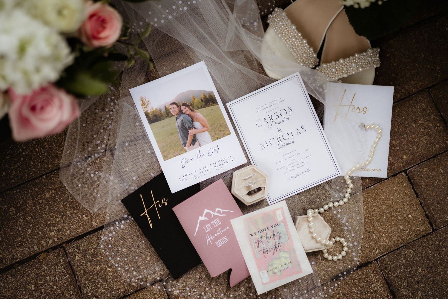 Elegant wedding invitation suite with save the date photo, floral cards, pearl necklace, and ring box on brick surface.