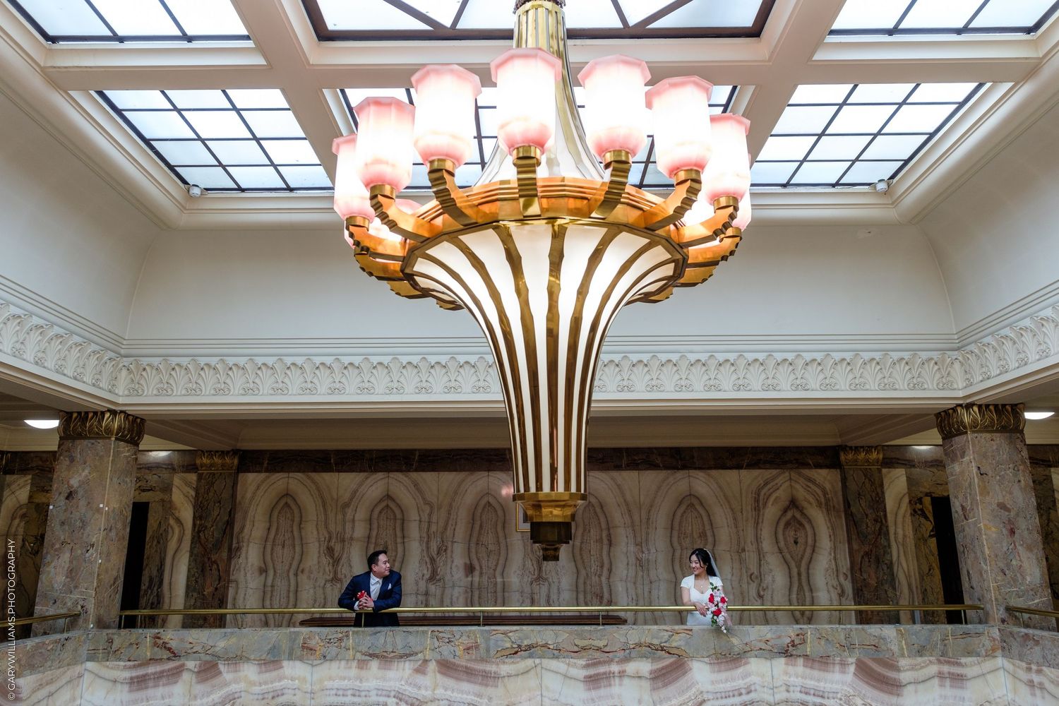 Elegant lobby with ornate gold chandelier, pink glass shades, marble walls, and two people standing on a mezzanine level.
