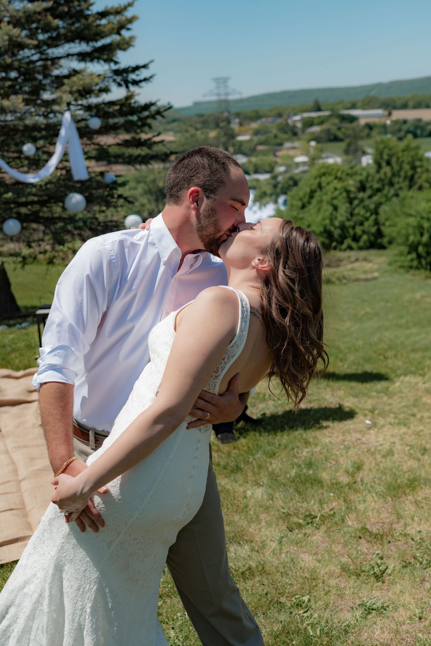 Bride and groom share a romantic kiss outdoors at a sunny summer wedding ceremony with green hills in the background.