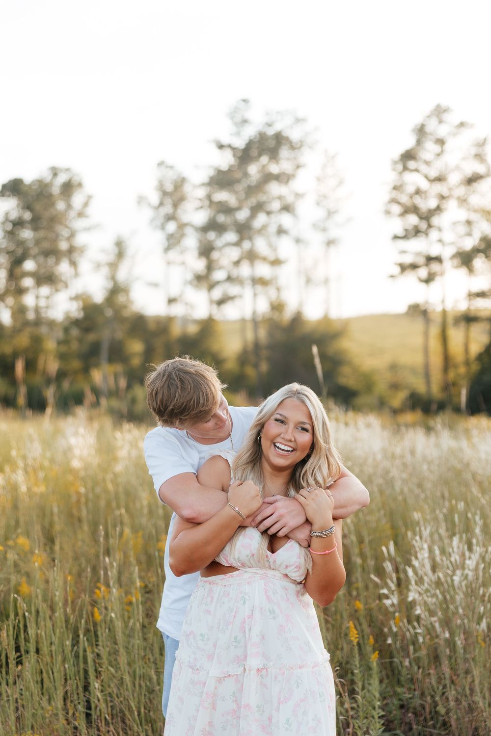 A laughing blonde woman in a floral dress embraced by a man in a sunny meadow with tall grass and trees.