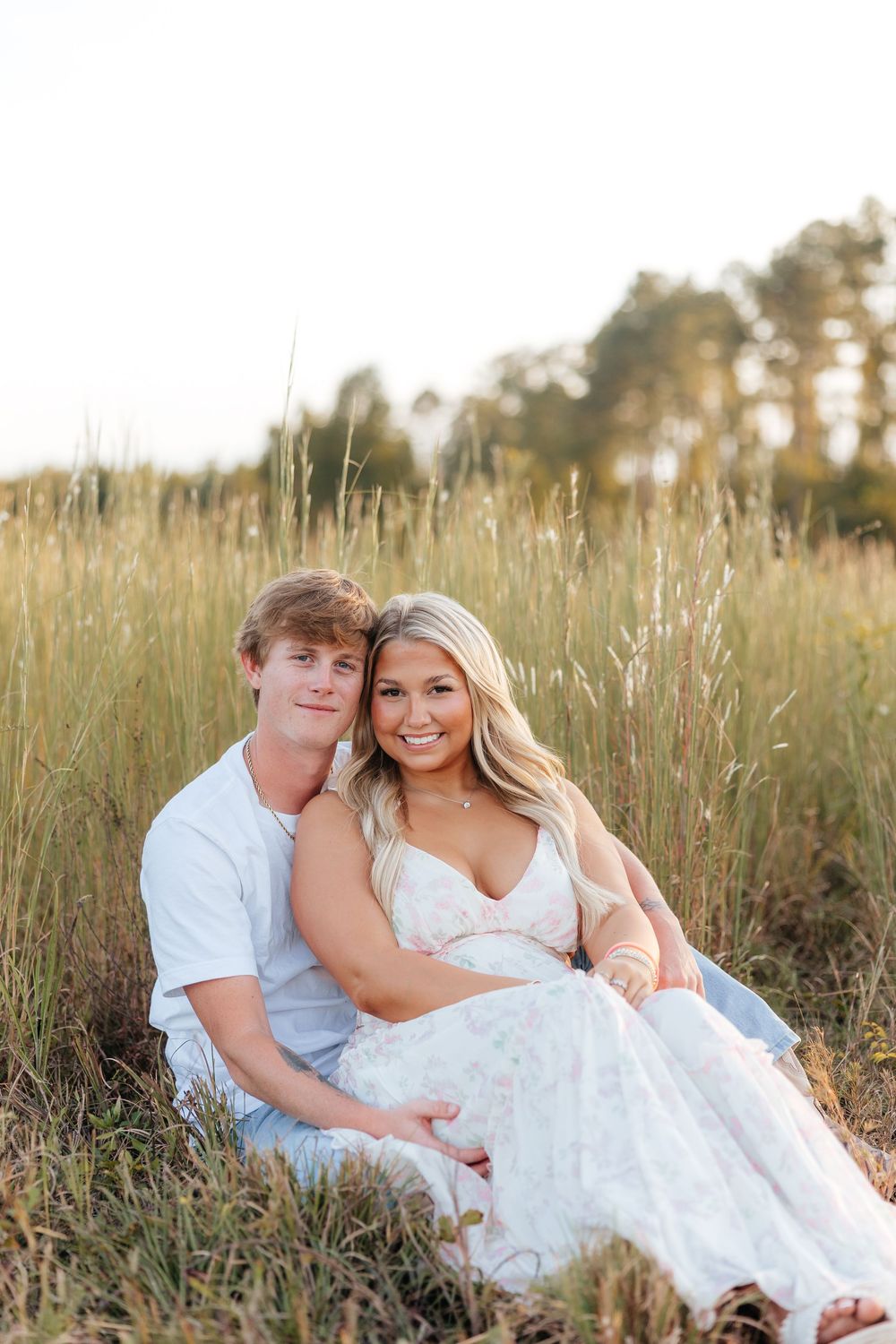A young couple sits together in a golden field of tall grass, smiling warmly at the camera during a sunny outdoor photo session.