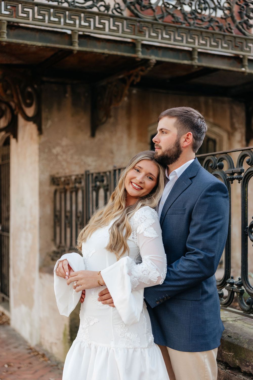 A couple embraces in front of an ornate iron fence, the woman in a white lace dress smiling as the man in a blue suit holds her.