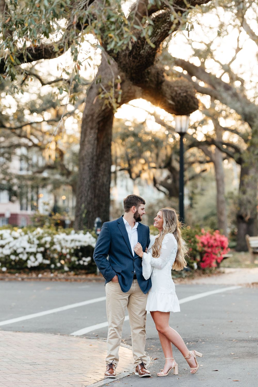 Couple in love standing under oak trees, man in navy blazer and woman in white dress, golden hour engagement photo.