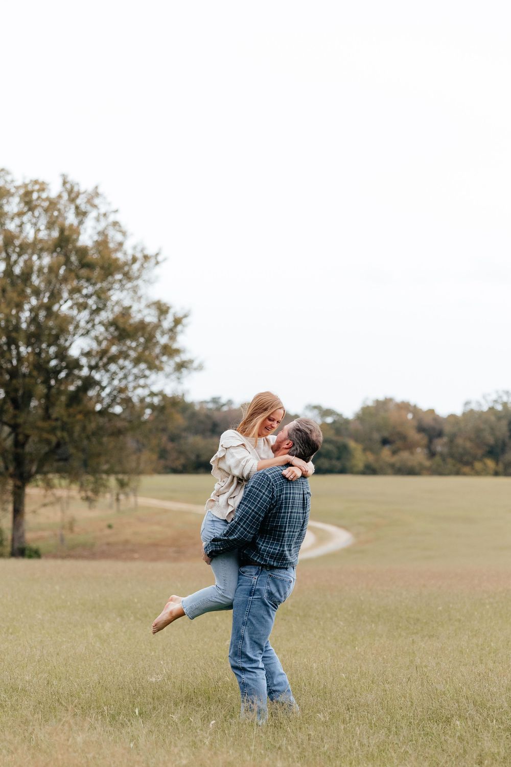 Couple embracing in a green field, man lifting woman as they share a romantic moment with trees in the background.