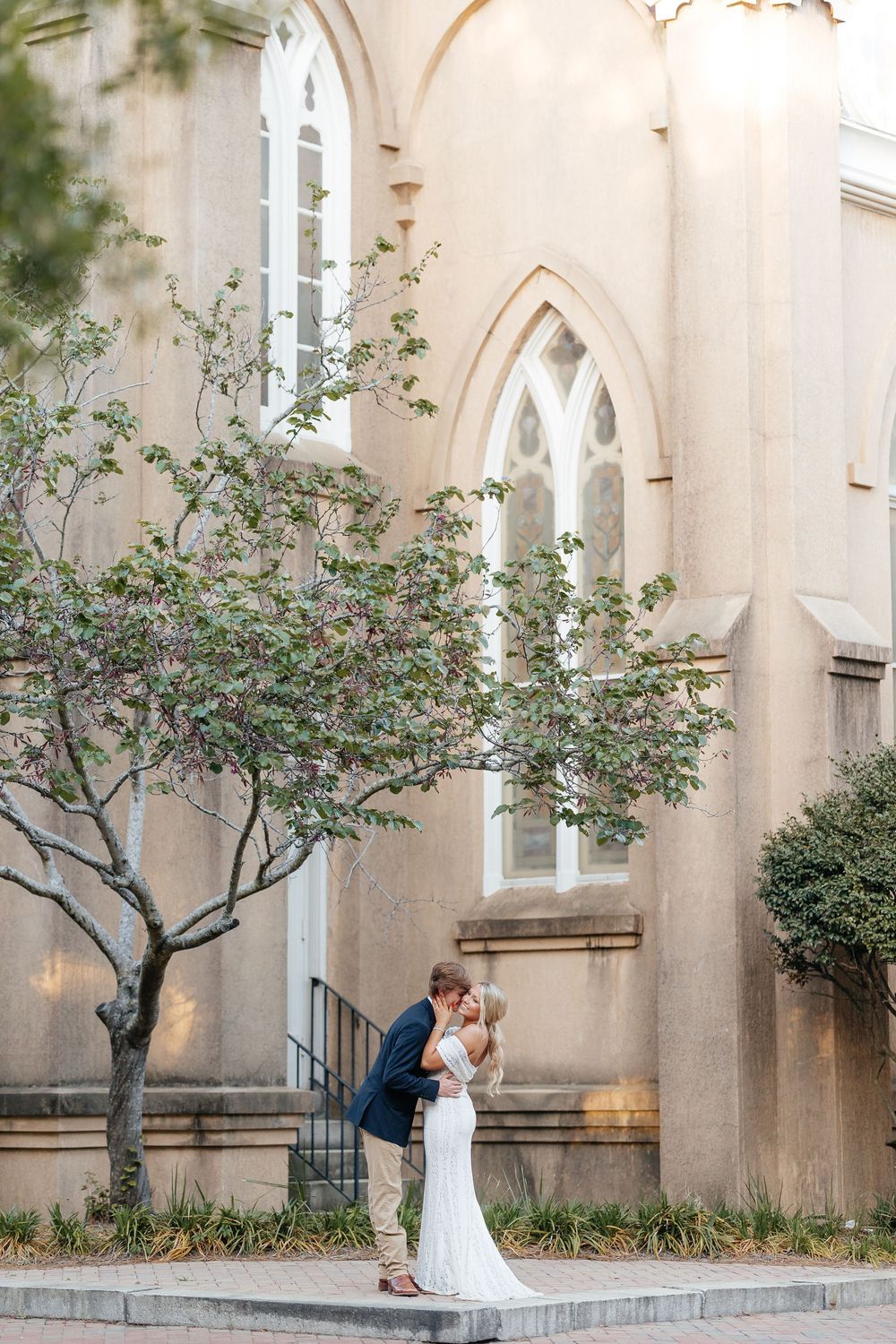 Bride and groom share a romantic kiss outside a historic stone church with Gothic arched windows and a blooming tree.