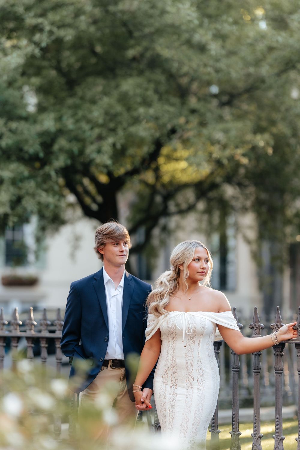 A couple in wedding attire holds hands by an iron fence, surrounded by lush greenery in an outdoor setting.