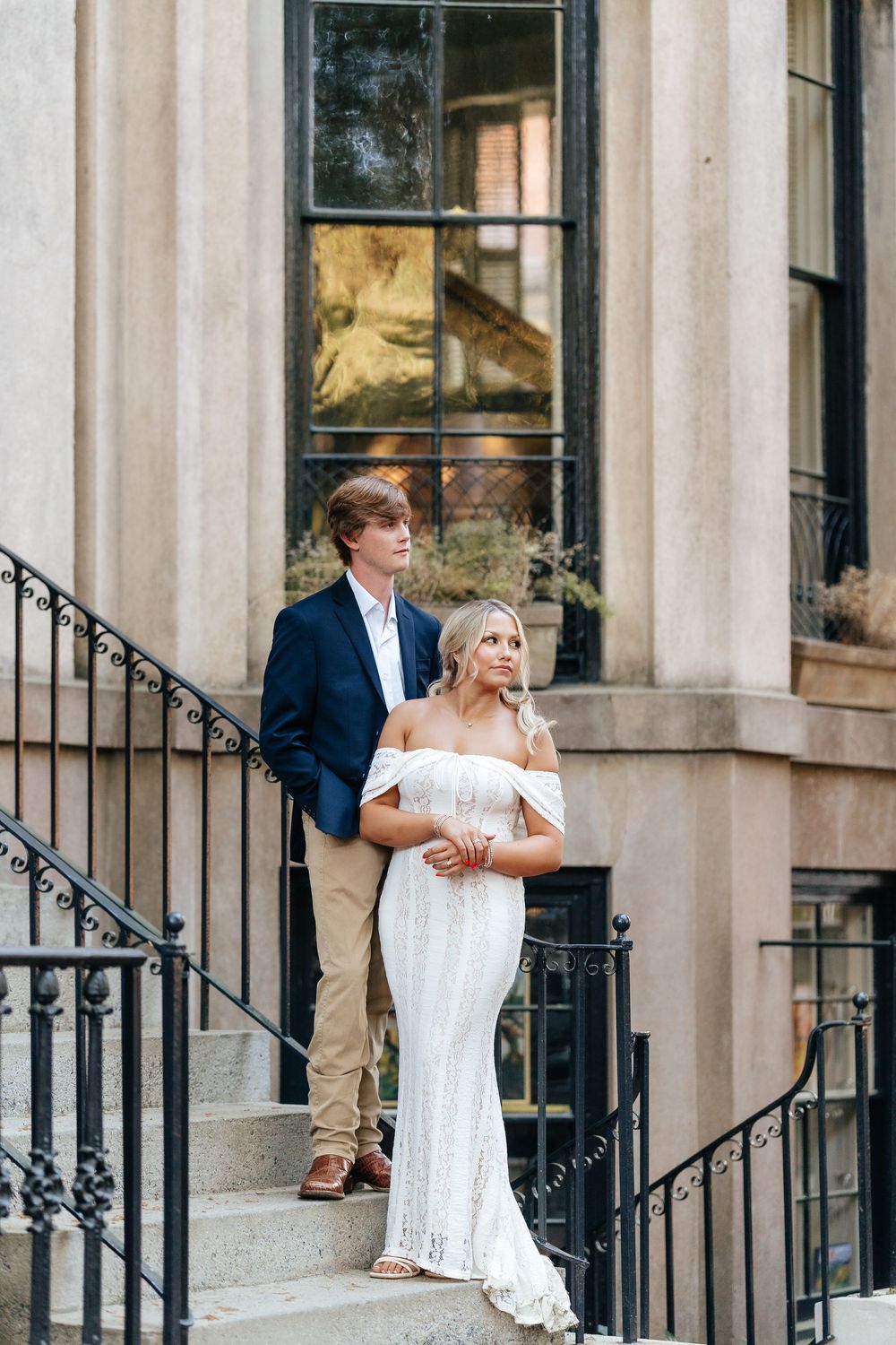 Elegant couple posing on stone steps, woman in white off-shoulder gown, man in navy blazer, beside classic building.
