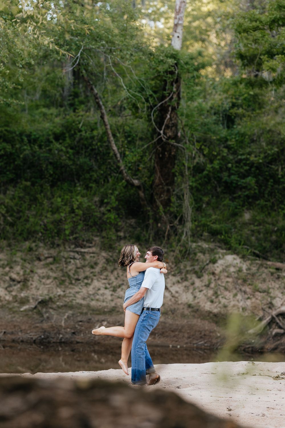 A couple embraces outdoors near a sandy riverbank surrounded by lush green trees.