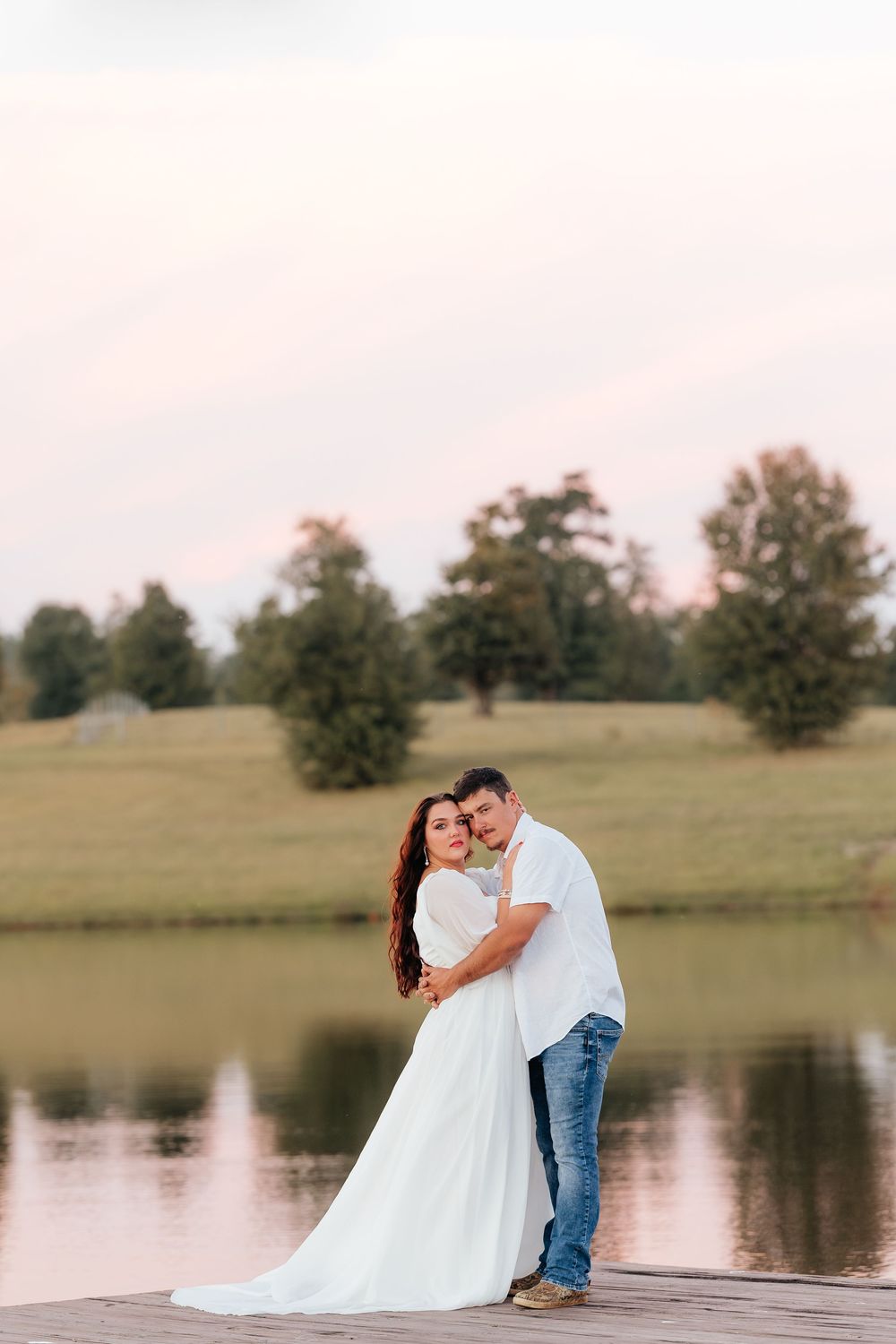 Couple embracing on a dock by a serene lake at sunset, woman in white gown, man in jeans and white shirt.