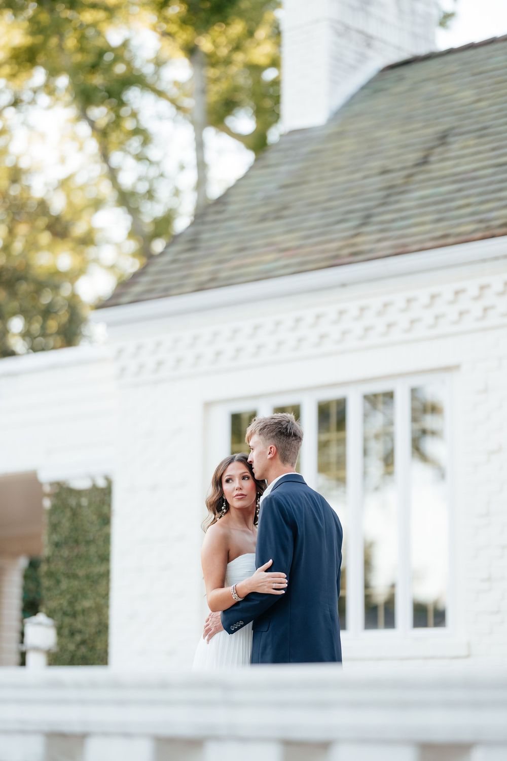 Bride in strapless white gown embraces groom in navy suit outside elegant white brick wedding venue.