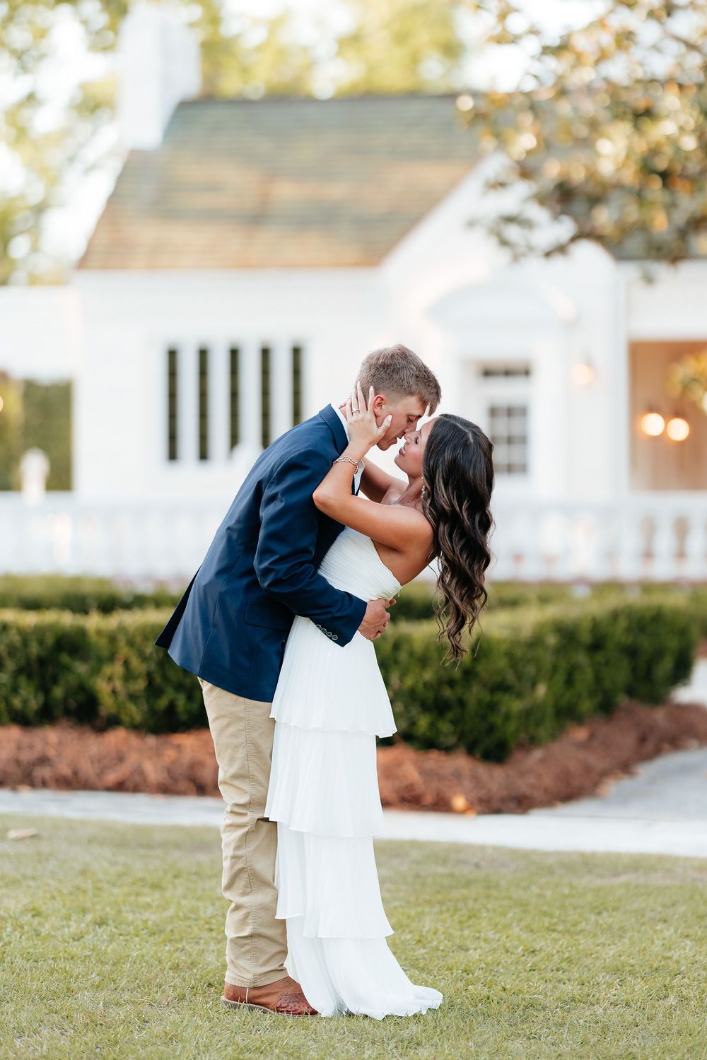 Bride and groom share a romantic dip kiss on green lawn before white chapel venue on their wedding day.