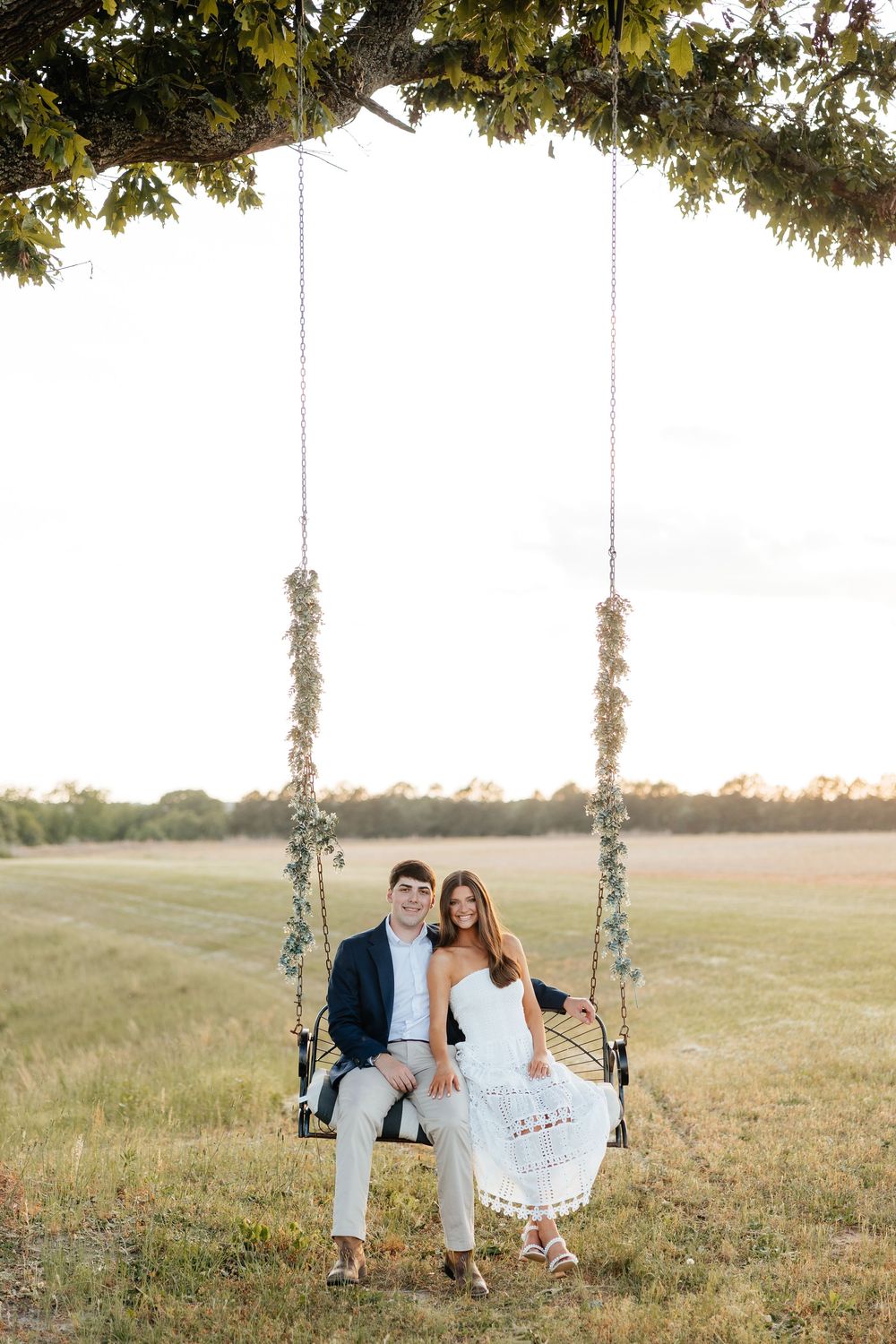 Couple sitting together on a decorated tree swing in a golden field during sunset engagement photo.