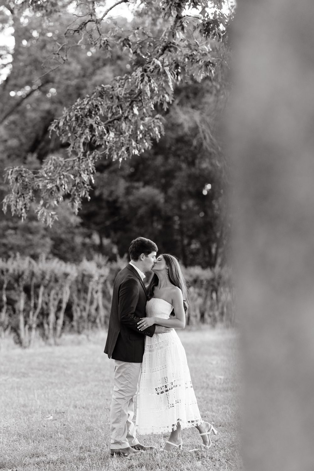 Black and white photo of a couple sharing a romantic kiss outdoors under a tree in a lush green garden.