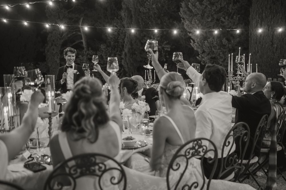 Wedding guests raise glasses in a toast at candlelit outdoor reception in black and white photo.